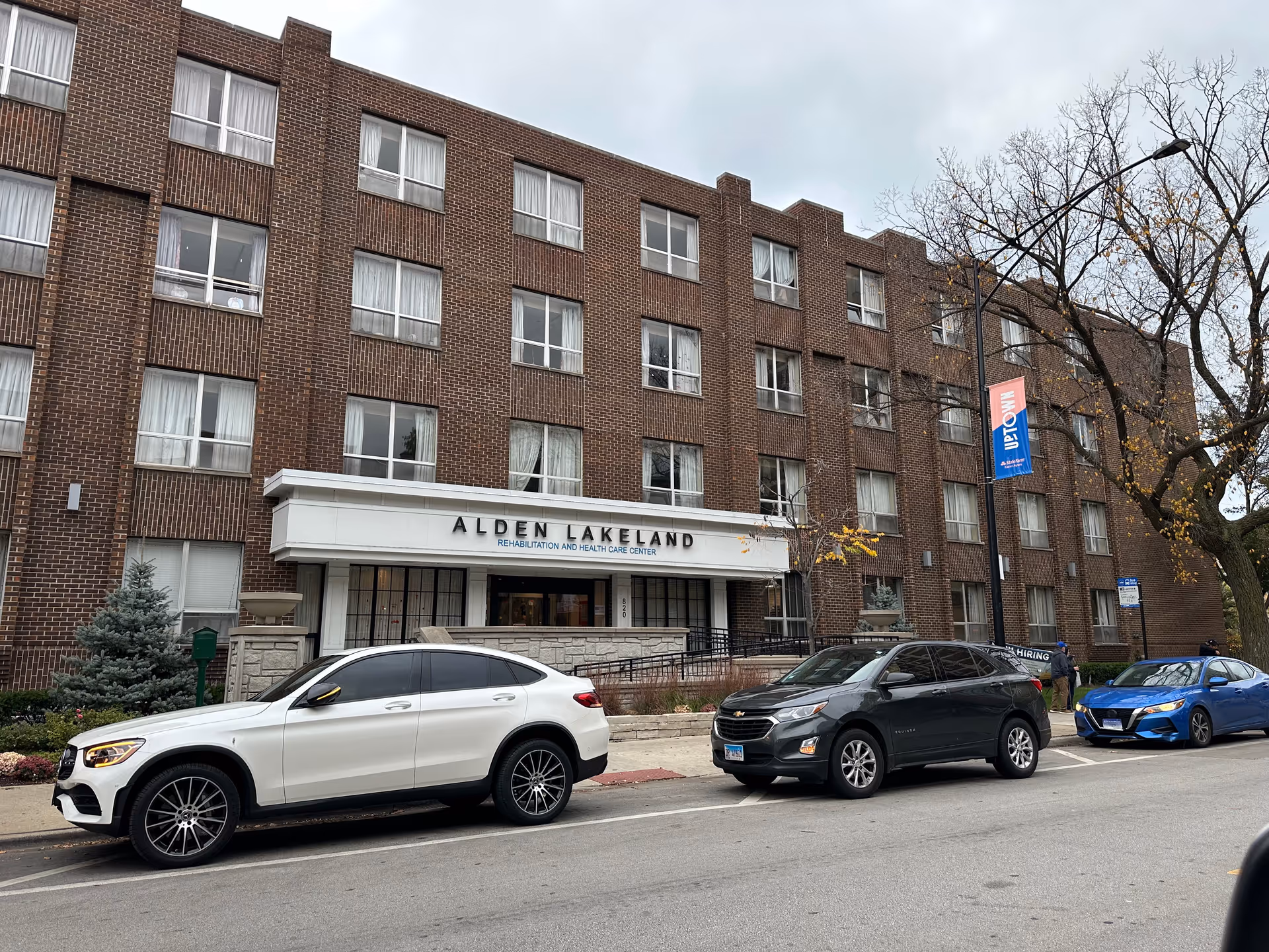 Exterior view of a multi-story brick building with a sign that reads 'Alden Lakeland Rehabilitation and Health Care Center'. Several cars are parked along the street in front of the building, and there is a tree with sparse leaves on the right side under a cloudy sky.