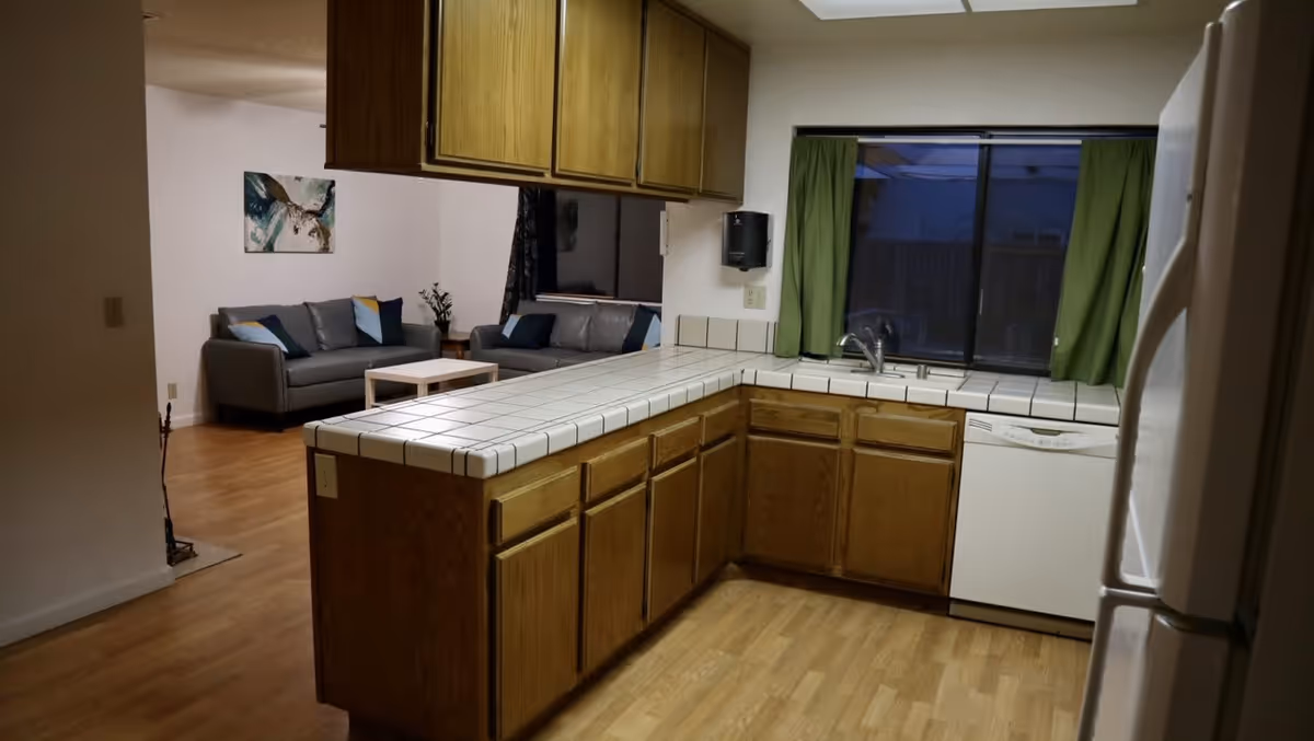 Interior view of a kitchen with wooden cabinets, white tiled countertops, a sink under a window with green curtains, a dishwasher, and a refrigerator. In the background, there is a living room area with two gray sofas, a small white coffee table, and a wall painting.