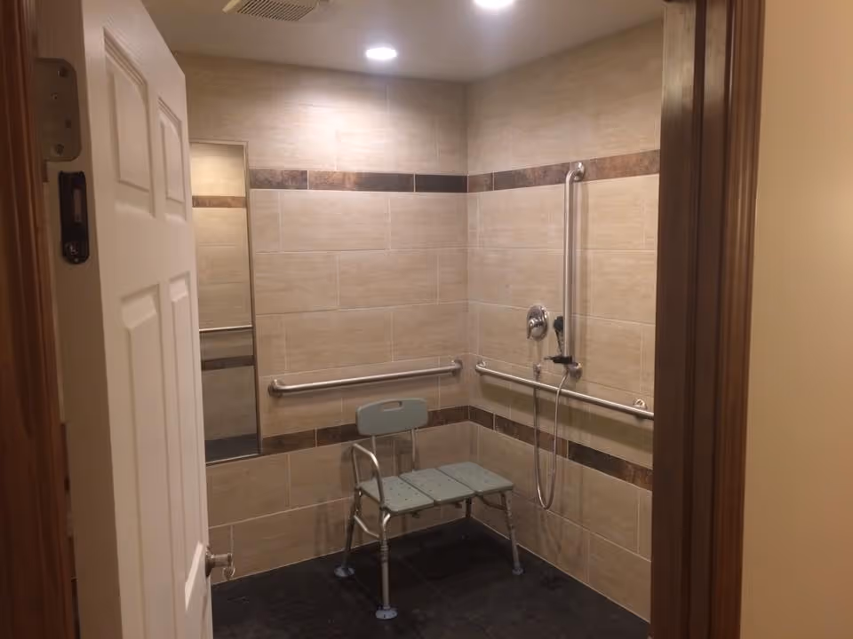 A tiled shower room with beige and brown tiles, equipped with metal grab bars on the walls and a green shower chair with backrest. There is a handheld showerhead attached to the wall and a mirror on the left side. The door to the room is partially open.