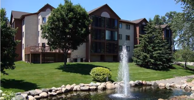 Exterior view of The Anthony James senior living facility with a fountain, pond, and green lawn.