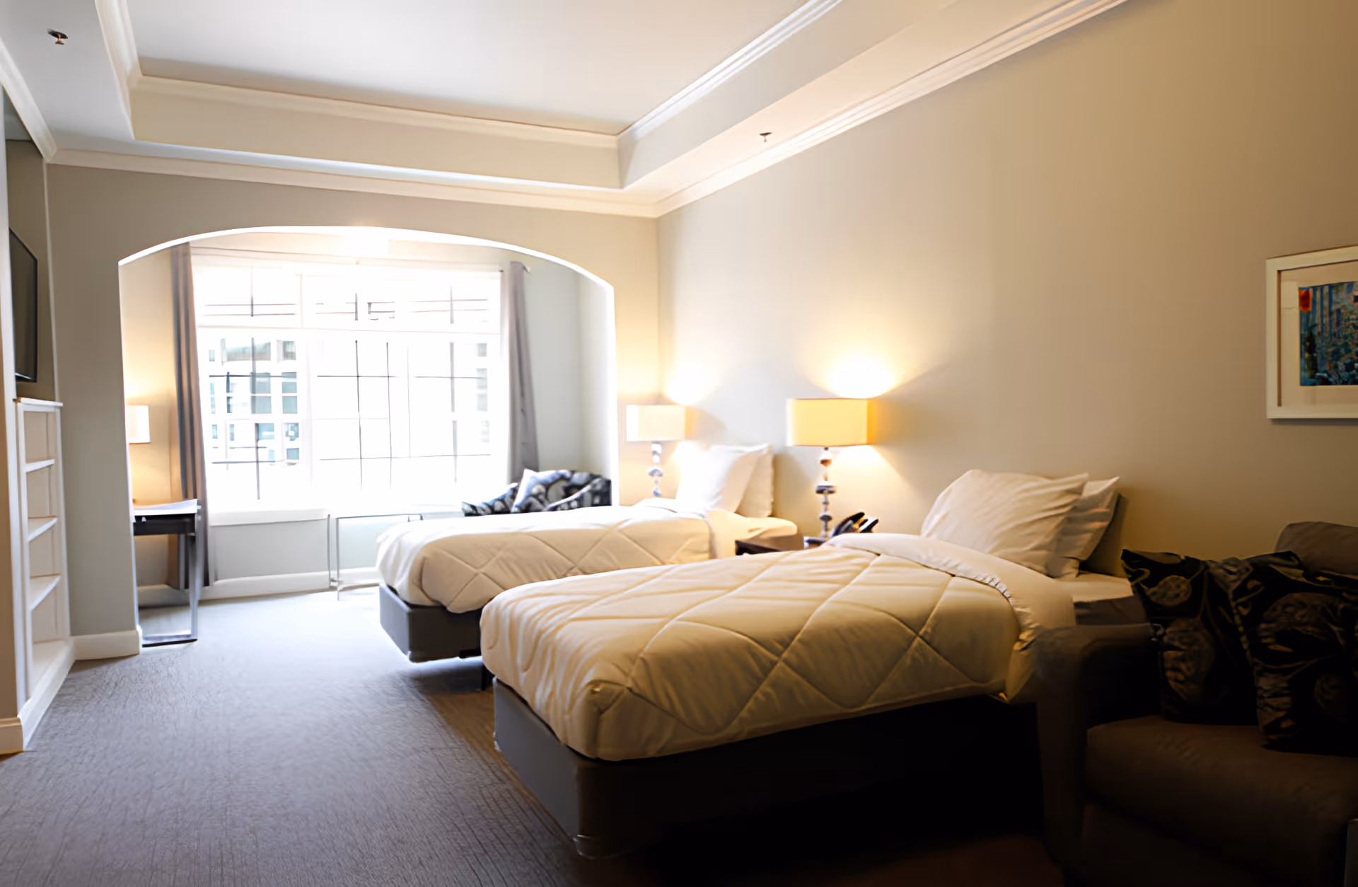 A bright and spacious bedroom with two single beds, each with white bedding and pillows. Between the beds is a nightstand with a lamp and a telephone. The room has a large window with curtains, a small desk, a wall-mounted TV, and a comfortable armchair with patterned cushions. The walls are painted light beige, and there is a framed picture hanging on the wall.