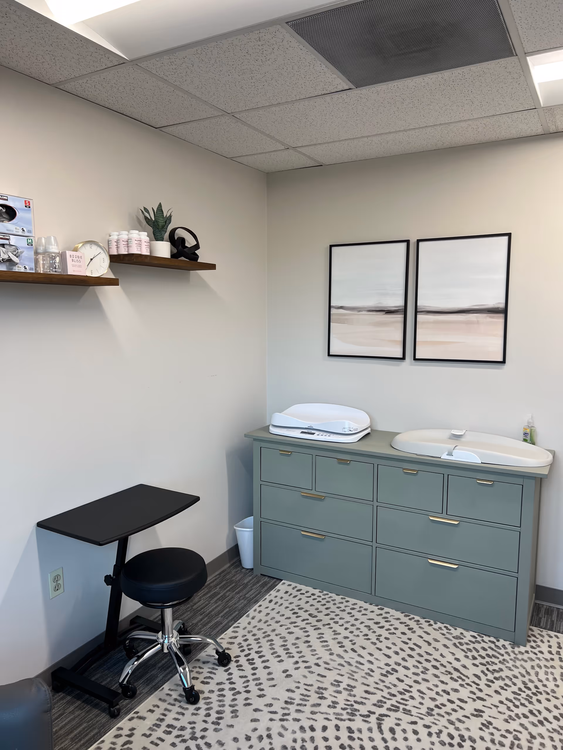 A clean lactation room with sink, mint-green cabinet, desk, stool, shelves, and wall art.