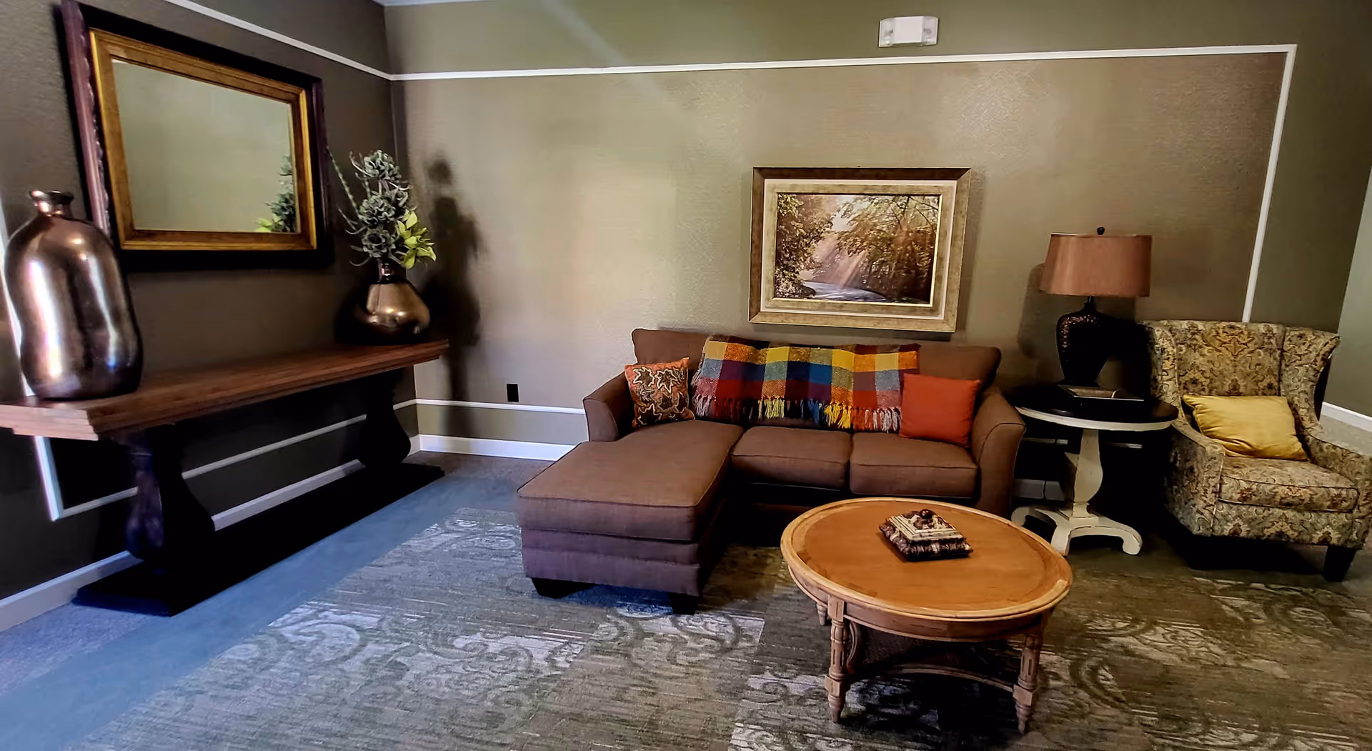 A cozy living room area with a brown sectional sofa adorned with colorful pillows and a plaid throw blanket. A round wooden coffee table is in front of the sofa, and a patterned armchair with a yellow pillow is to the right. On the left side, there is a wooden console table with decorative vases and a large mirror above it. A framed picture of a forest scene hangs on the wall above the sofa. The room has neutral-colored walls and carpeted flooring.
