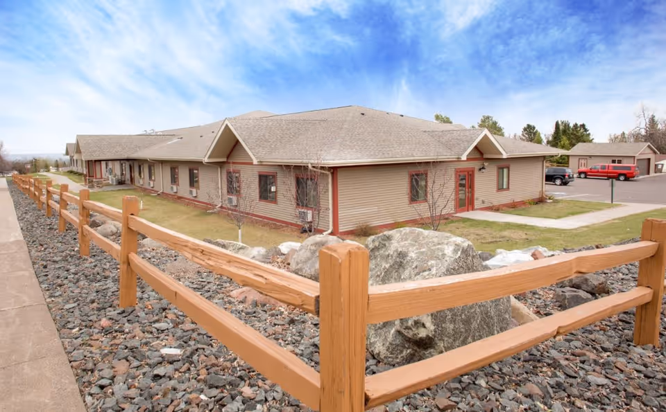 Single-story beige senior living facility with a wooden fence and rocky landscaping.