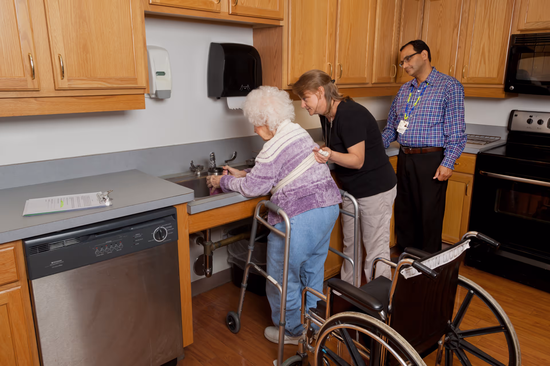 An elderly woman using a walker is washing her hands at a kitchen sink with assistance from a caregiver, while a man stands nearby observing. The kitchen has wooden cabinets, a dishwasher, and a stove, with a wheelchair positioned close to the woman.