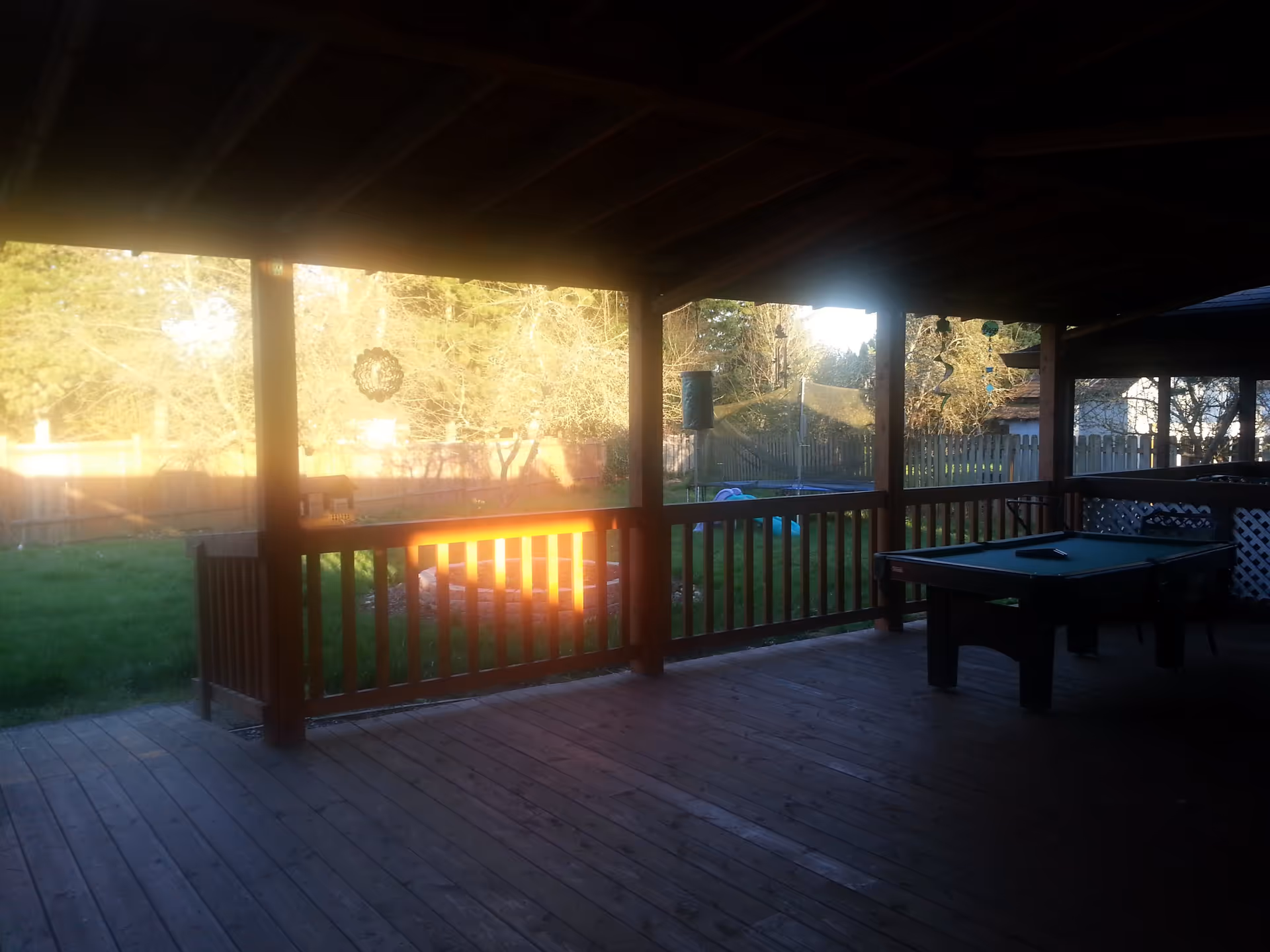 Covered wooden patio with a pool table, overlooking a fenced backyard with green grass and trees in the background during daylight.