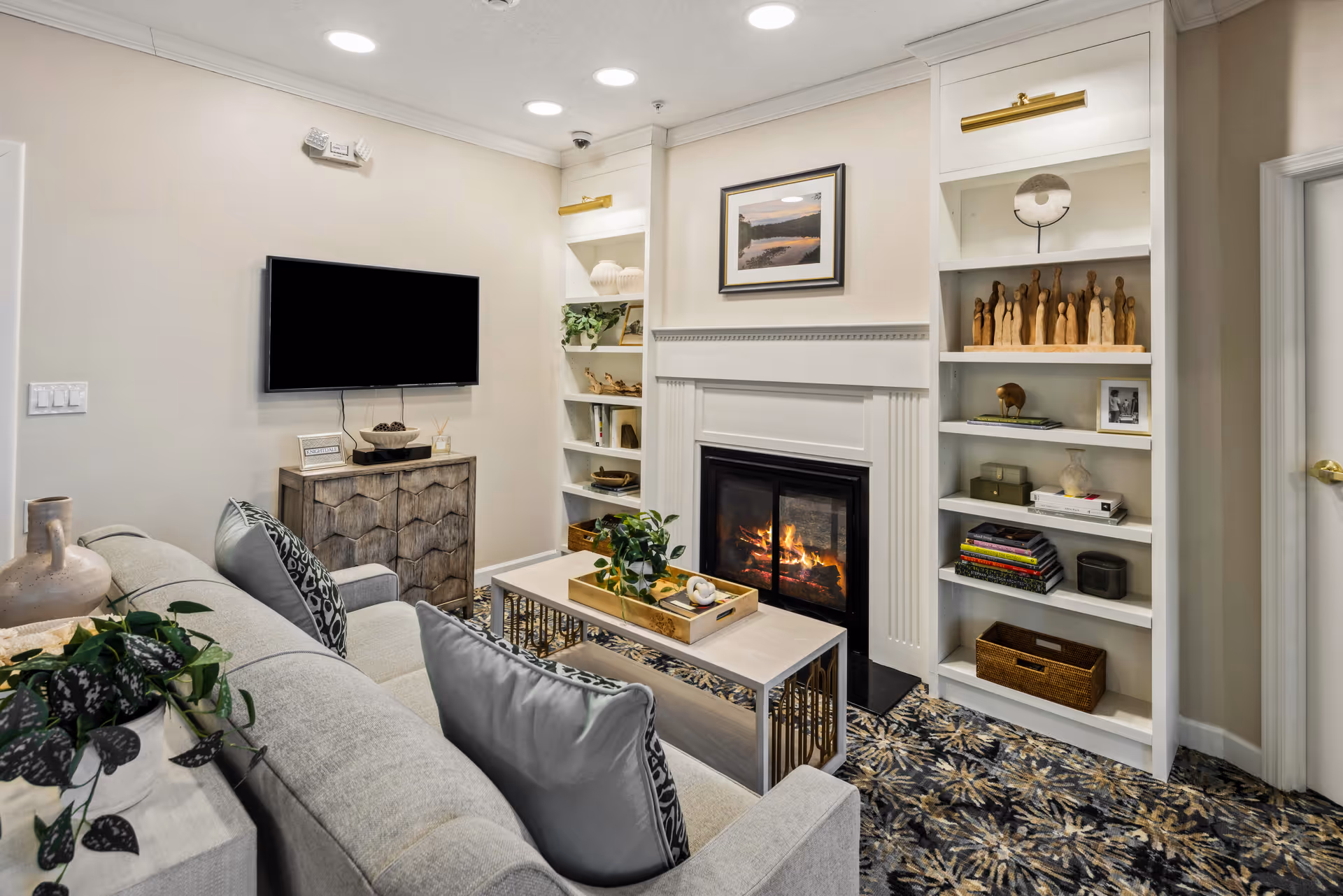 A cozy living room with a light gray sofa adorned with patterned pillows, a coffee table with a decorative tray and plant, a wall-mounted flat screen TV above a wooden cabinet, built-in white shelves with decorative items, and a fireplace with a fire burning. The room has a patterned carpet and neutral-colored walls.
