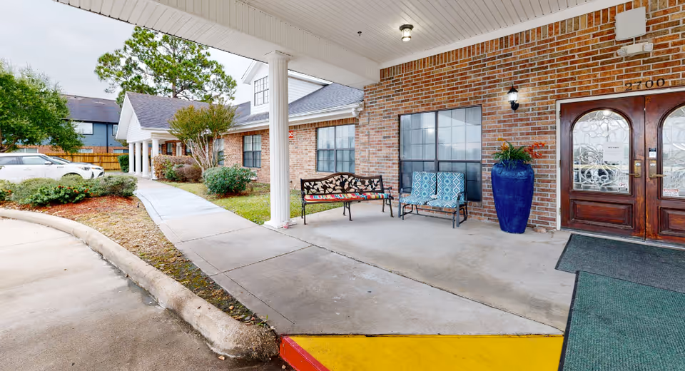 Covered entrance to a brick senior living building with benches, a blue planter, and double doors.