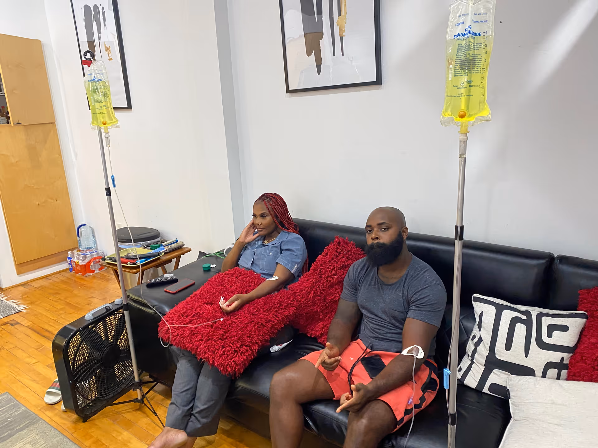 Two adults sit on a black leather sofa in a living room with IV stands.