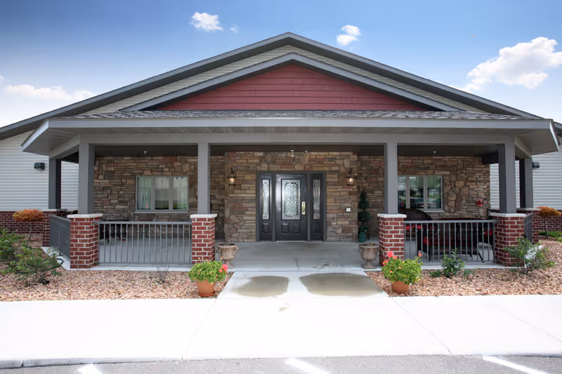 Front exterior view of a single-story building with a covered porch supported by columns. The facade features stone and brick accents, with a central double door entrance and windows on either side. There are potted plants and landscaping around the porch area under a clear blue sky.