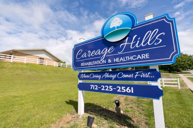 A large blue sign for Careage Hills Rehabilitation & Healthcare on a grassy area with a white fence and a building in the background under a partly cloudy sky.