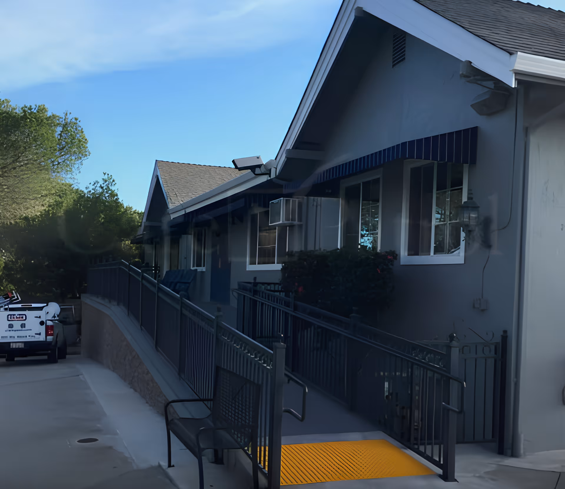 Exterior view of a gray assisted living building with a wheelchair ramp, bench, windows, and a parked truck.