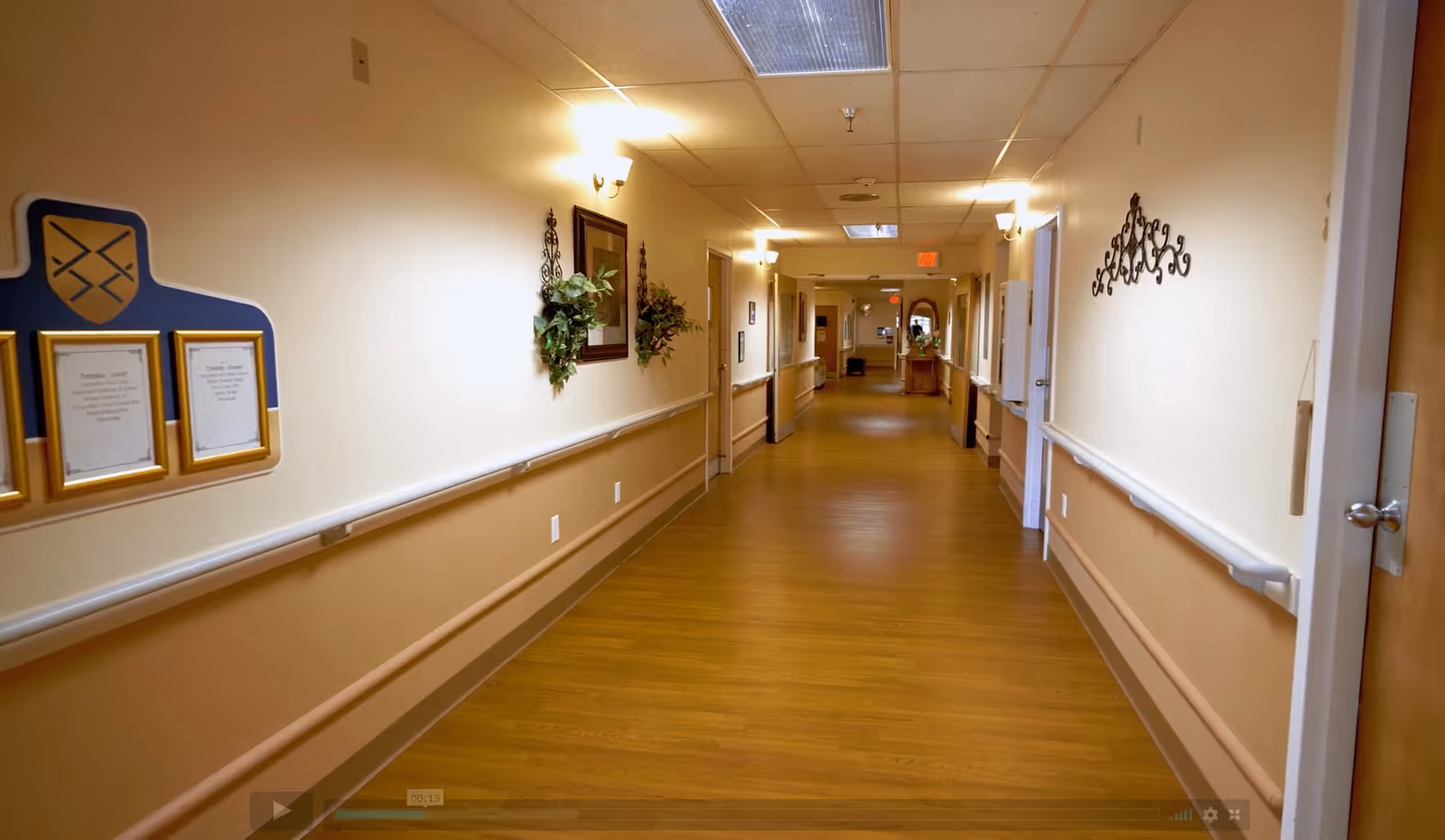 A long, well-lit hallway in a senior living facility with wooden flooring and beige walls. The hallway has handrails on both sides, wall-mounted lights, framed pictures, and decorative plants. Several doors line the hallway, and there is a small table with a mirror at the far end.
