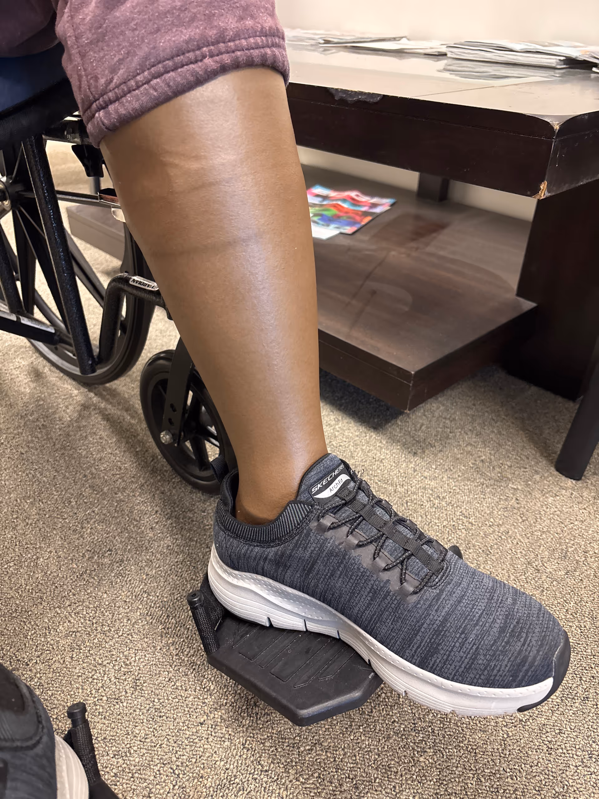 Close-up of a leg in a gray-blue sneaker beside a wheelchair and desk.