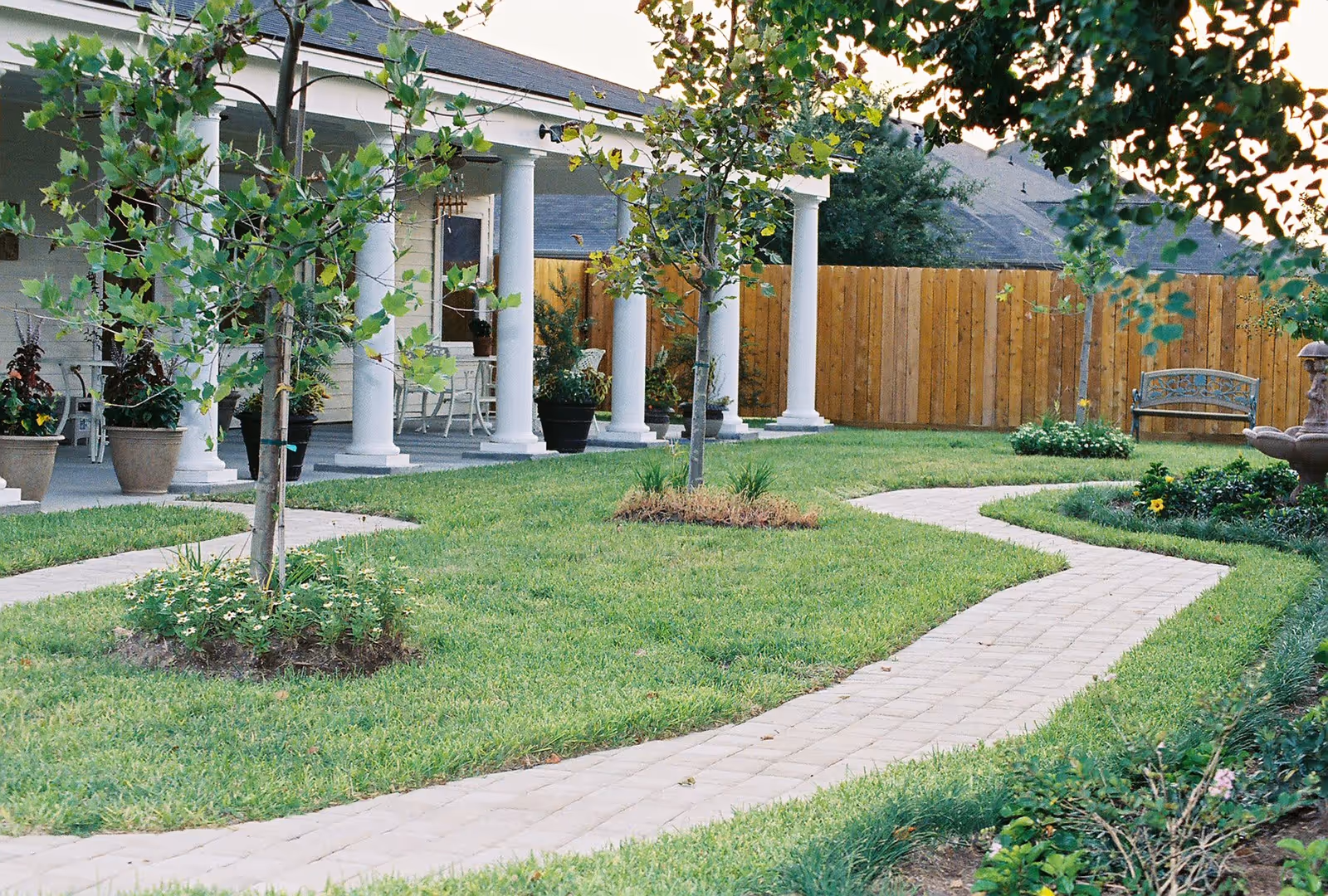 A landscaped outdoor garden area with a winding paved walkway, green grass, small trees, flower beds, and a wooden fence. There is a covered porch with white columns and outdoor seating visible in the background.
