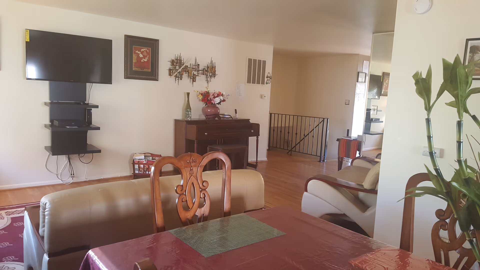 A spacious senior living lounge with a dining table, couch, piano, and wall-mounted TV.