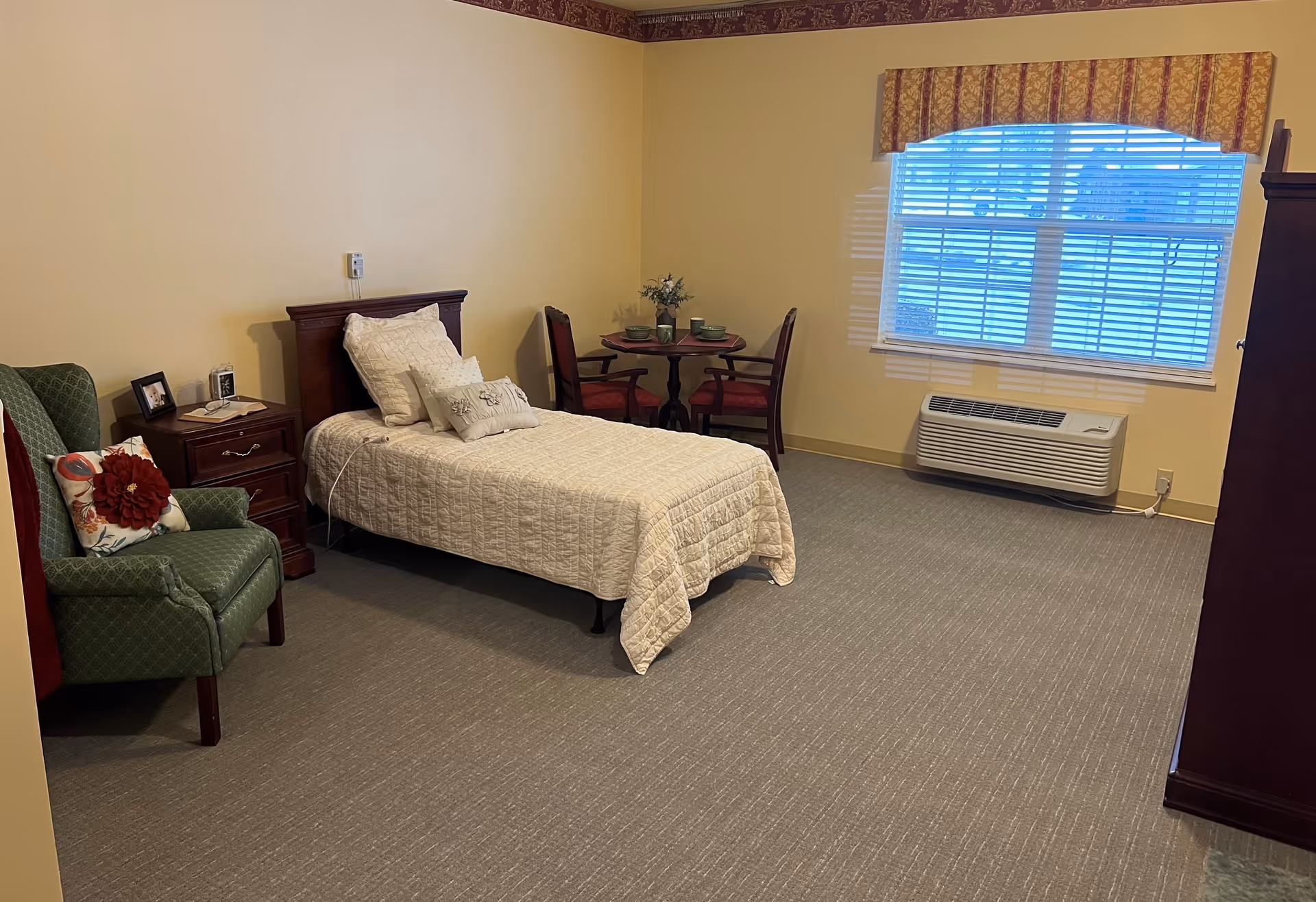 Furnished senior-living bedroom with a single bed, nightstand, upholstered chair, small dining table, and a window air-conditioning unit.