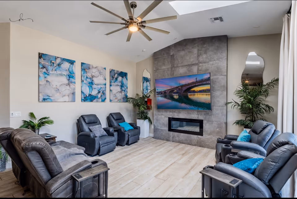 Cozy senior living lounge with recliners, a TV, art, plants, and a ceiling fan.