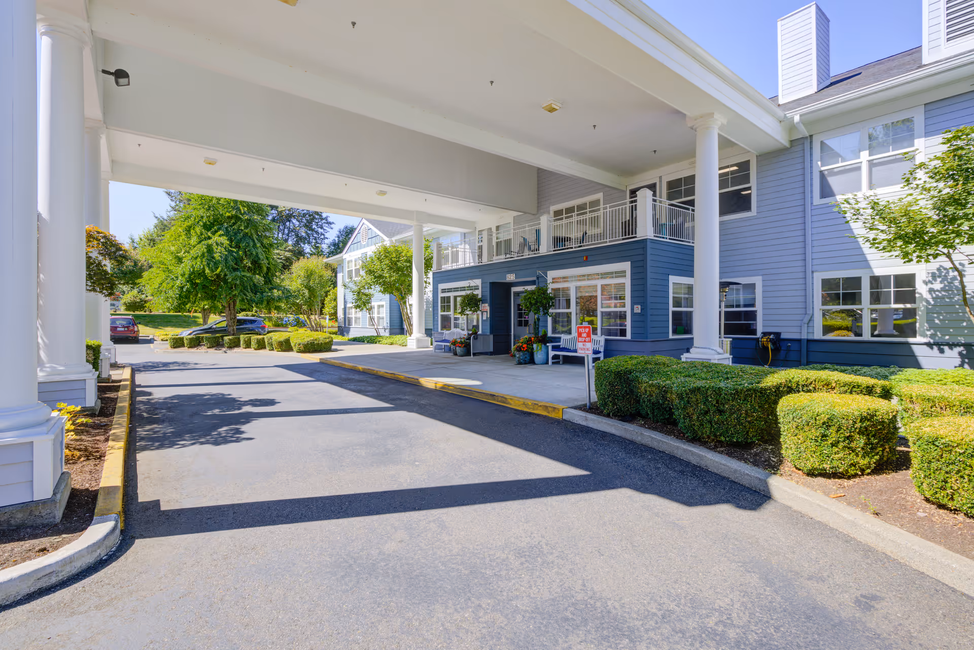 Covered driveway leads to the front entrance of a blue assisted living building with shrubs.
