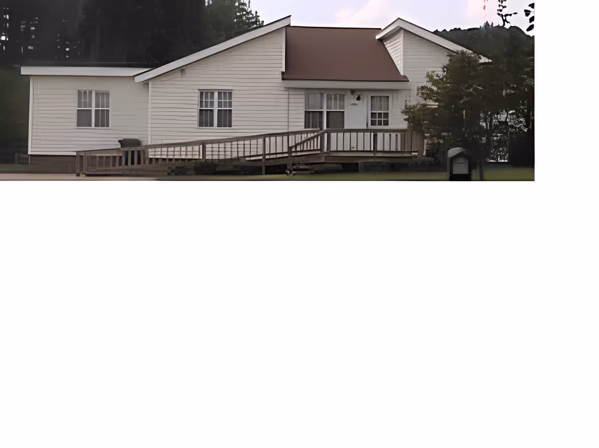 Exterior view of a single-story white building with a brown roof and a wooden ramp leading to the entrance, surrounded by grass and trees.