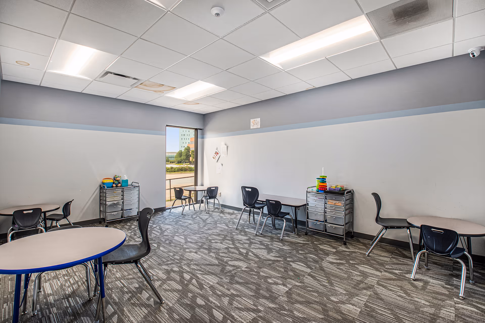 An interior activity room with round and rectangular tables, chairs, and storage carts.