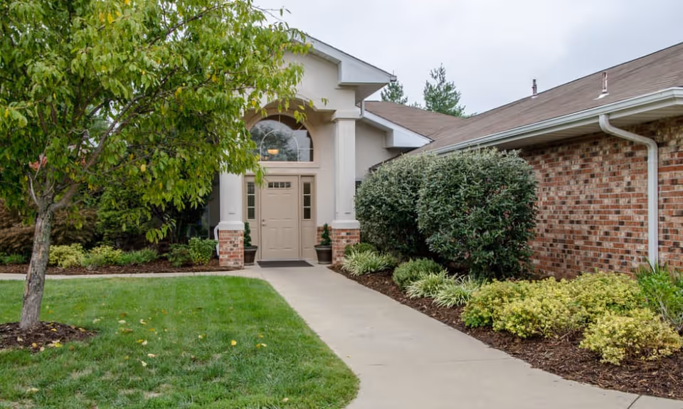 Exterior view of a senior living facility entrance with a concrete walkway leading to a beige double door. The building has a brick and stucco facade with a pitched roof. There are green shrubs and a tree on either side of the walkway, along with well-maintained landscaping and grass.