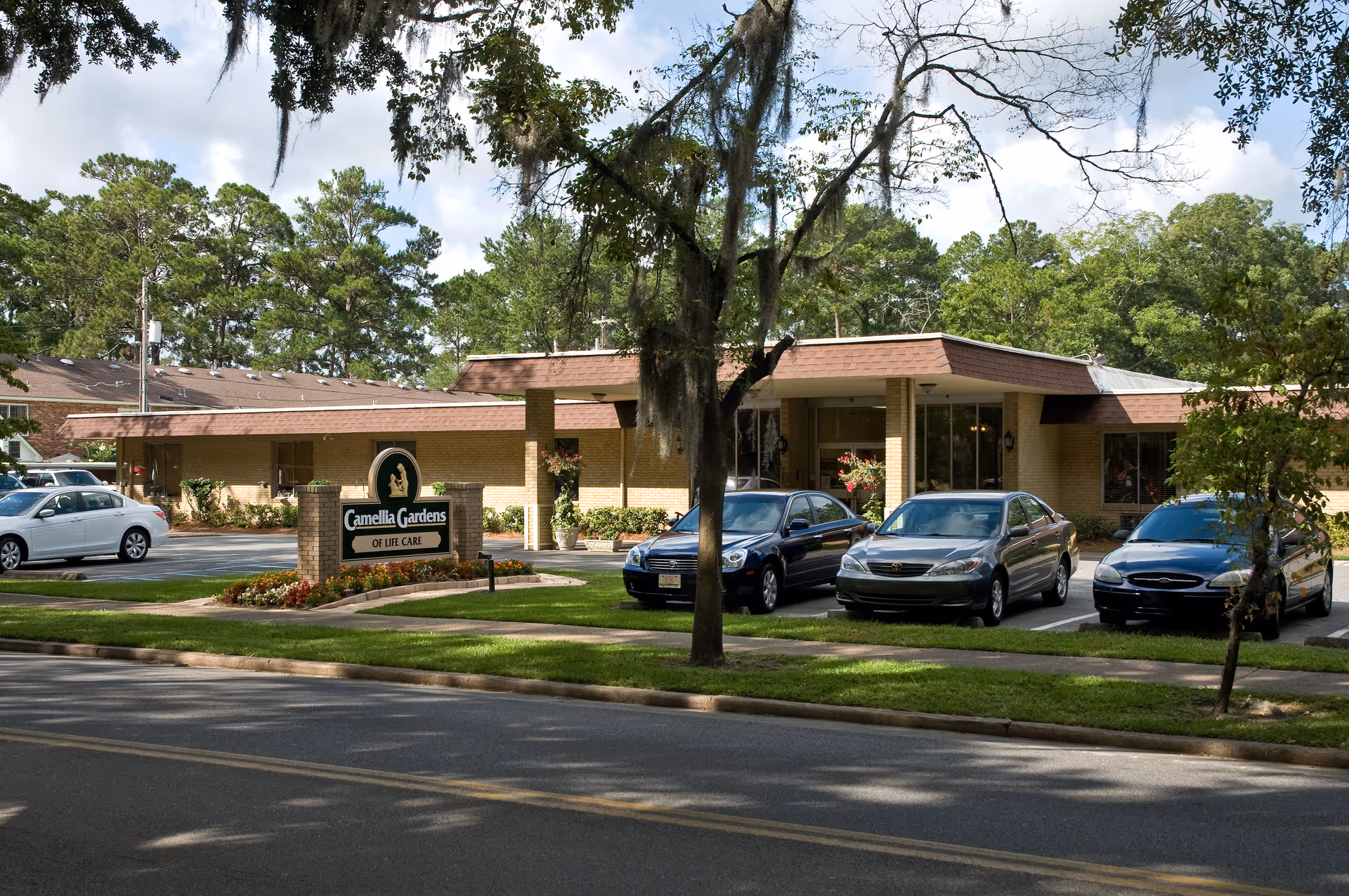 Exterior view of Camellia Gardens of Life Care facility showing a single-story brick building with a brown roof, several parked cars in front, a landscaped area with flowers, and a sign displaying the facility's name. Trees and greenery surround the building under a partly cloudy sky.