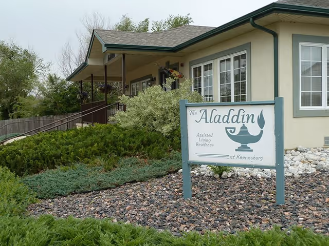 Exterior view of The Aladdin Assisted Living building with a sign and landscaped front yard.