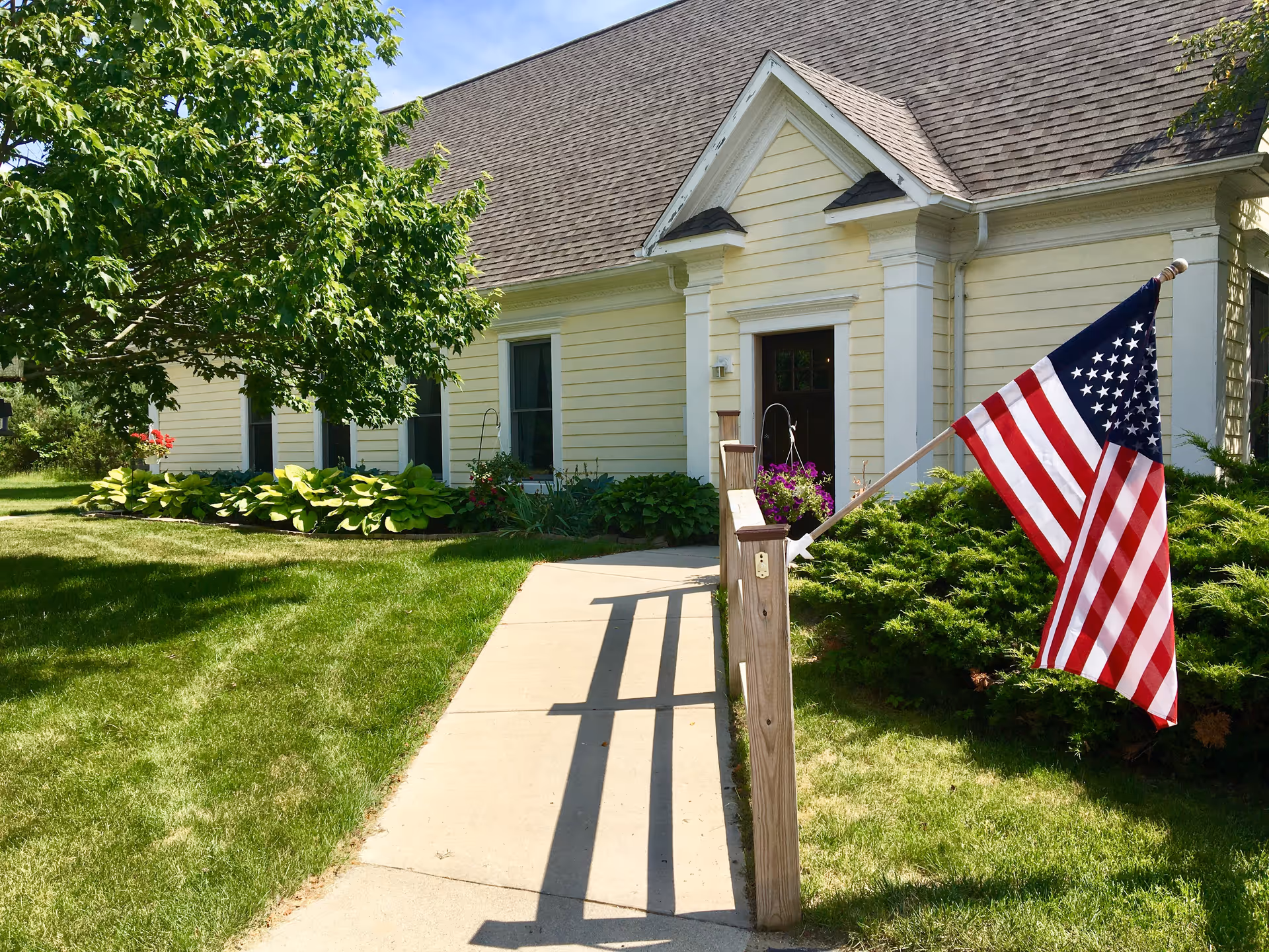A yellow single-story house with white trim and a brown shingled roof. A concrete walkway leads to the front door, with a wooden handrail on one side displaying an American flag. The yard is well-maintained with green grass, bushes, and a tree providing shade.