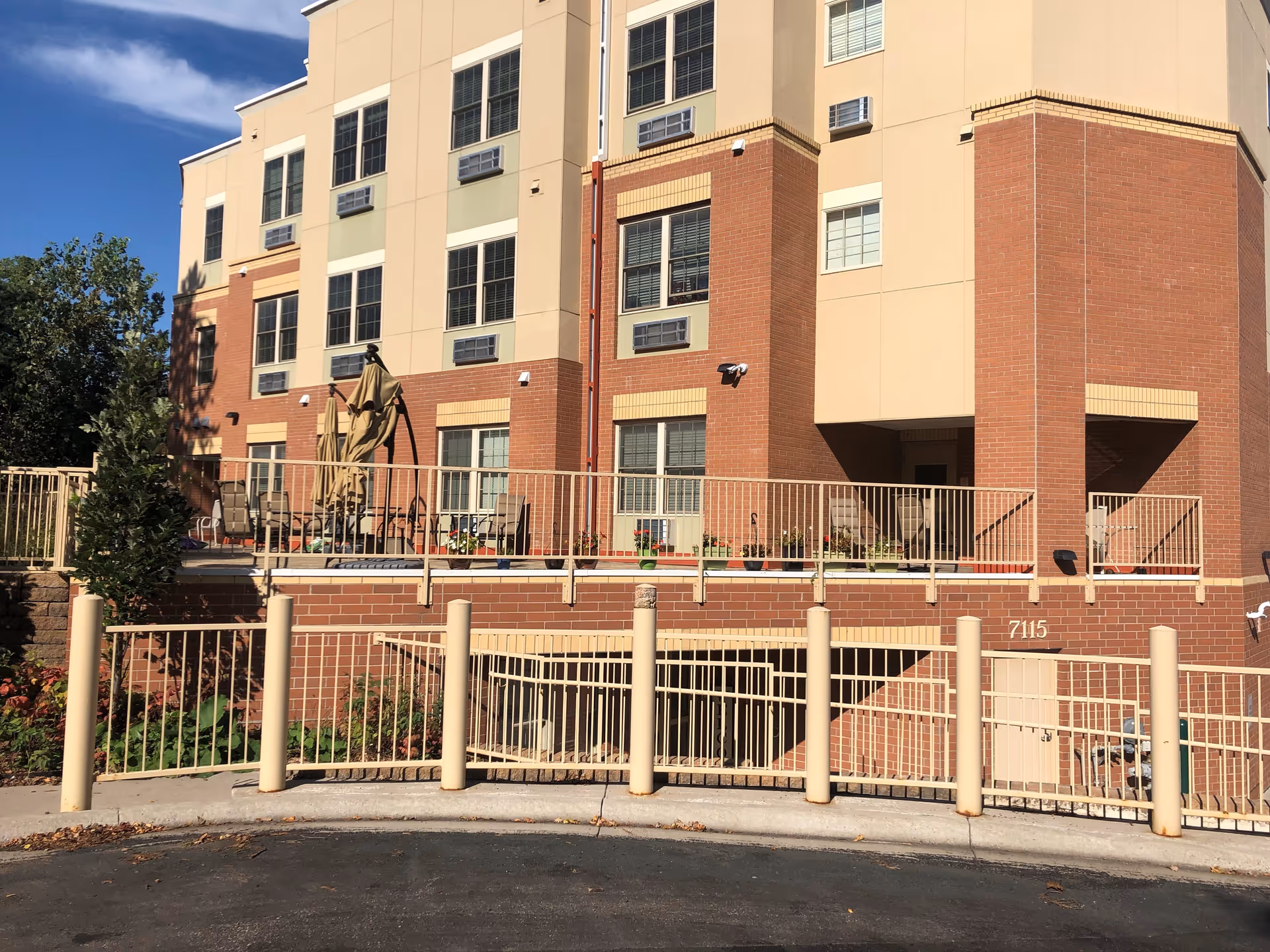 Exterior view of a multi-story senior living facility building with beige and red brick walls, multiple windows, and a fenced patio area with outdoor furniture and umbrellas. The building number 7115 is visible on the brick wall near the entrance.