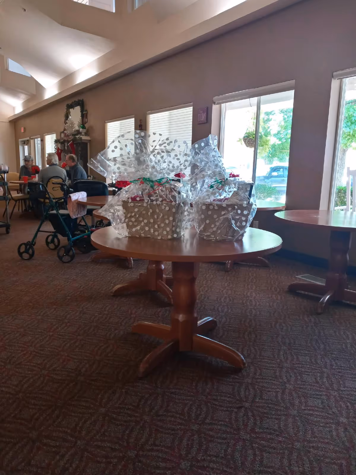 Interior senior living common area with round tables, gift baskets, and residents seated nearby.
