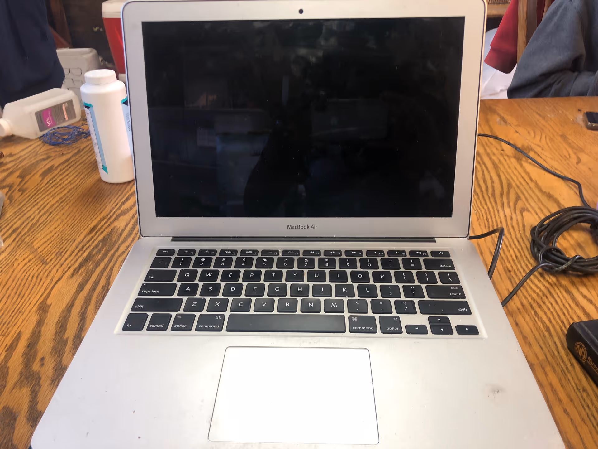 Open silver MacBook Air on a wooden table with cables and assorted supplies nearby.