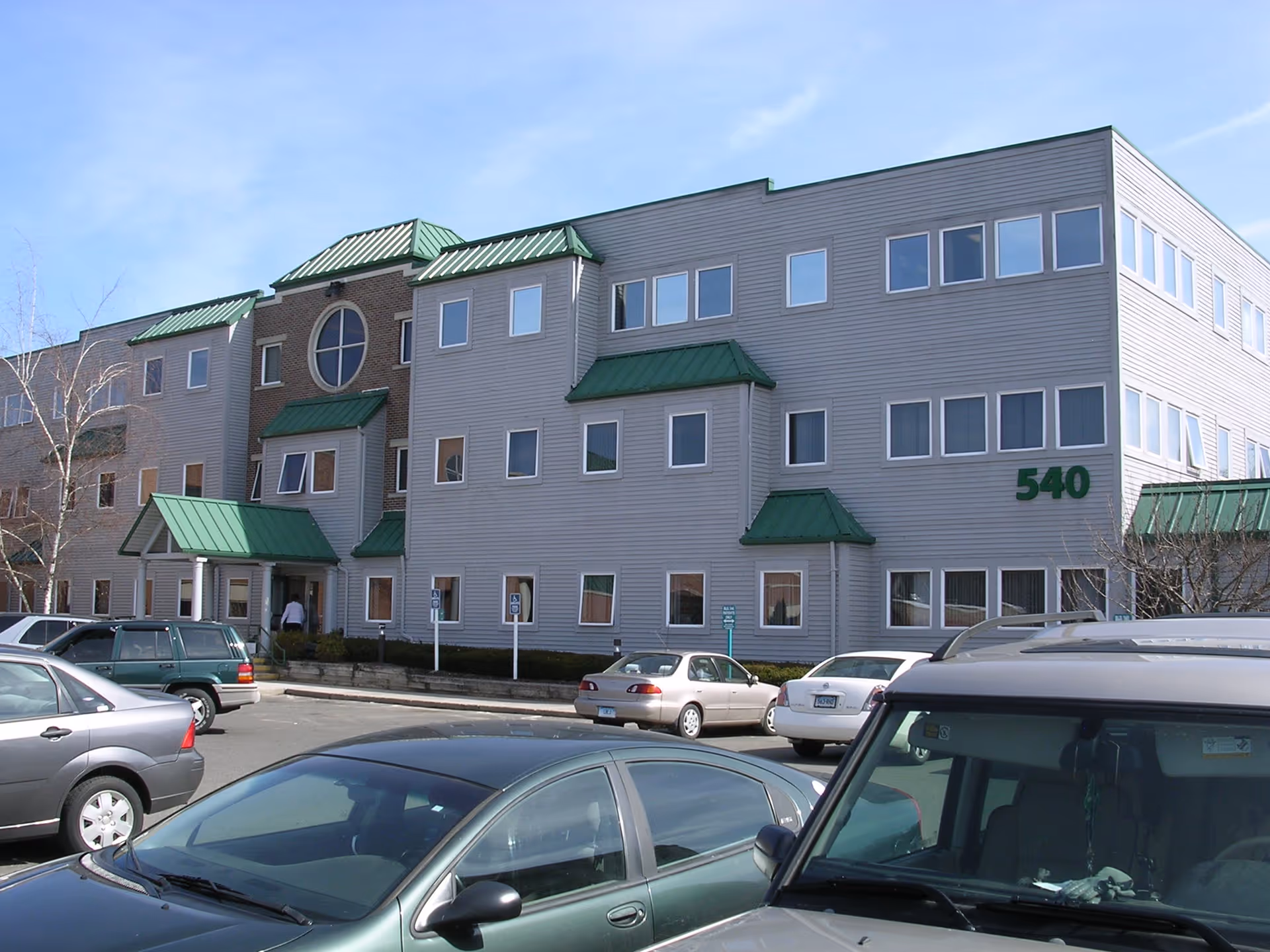 Gray multi-story building with green roofs and parked cars in front.