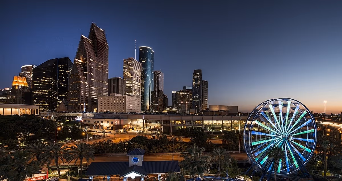 Nighttime city skyline with tall buildings and a brightly lit Ferris wheel.
