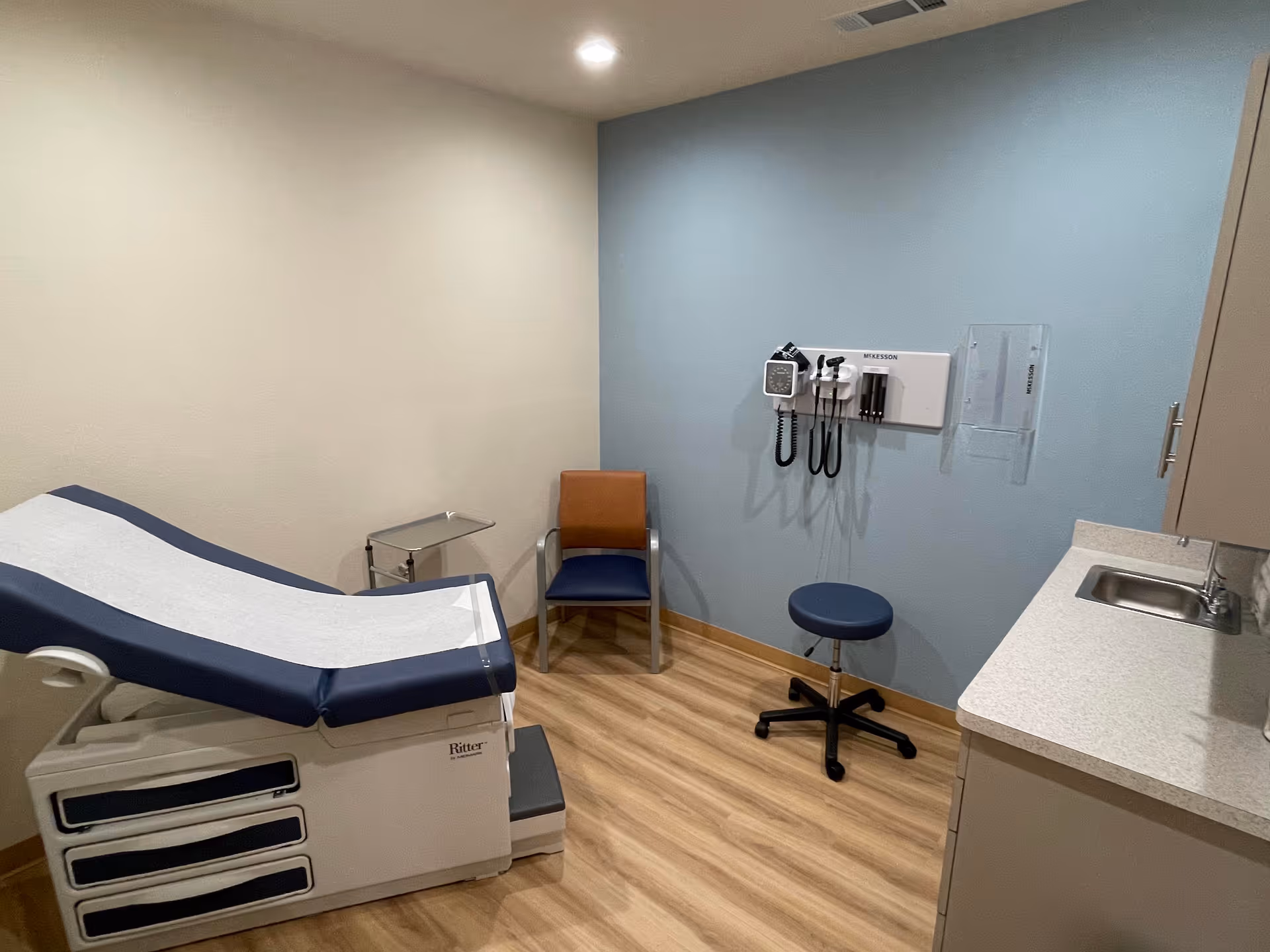 Interior exam room with a reclining medical bed, chair, and mounted diagnostic equipment.