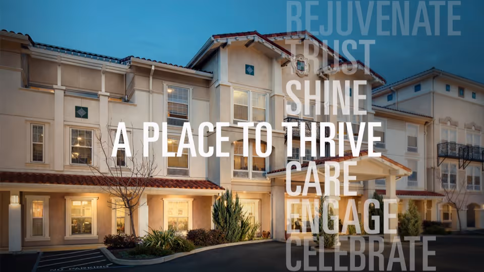 Exterior view of Belmont Village Senior Living Sunnyvale building at dusk with warm lights glowing from the windows, a driveway, and some landscaping including small trees and bushes.