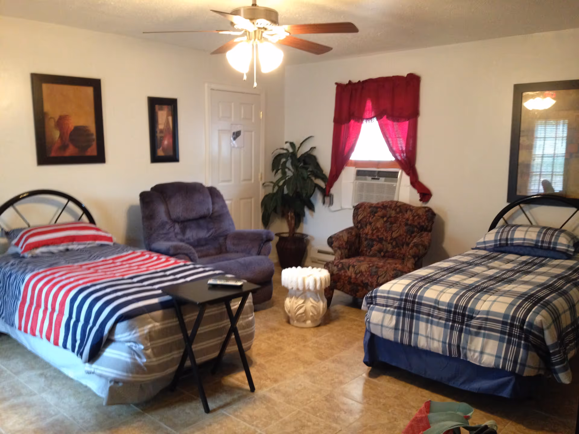 A bedroom with two single beds, one with red, white, and blue striped bedding and the other with blue plaid bedding. Between the beds is a small black folding table with a remote control on it. There are two armchairs, one purple and one floral patterned, with a decorative white stool between them. A potted plant is in the corner near a window with red curtains and an air conditioning unit. The walls have framed artwork, and a ceiling fan with lights is overhead.