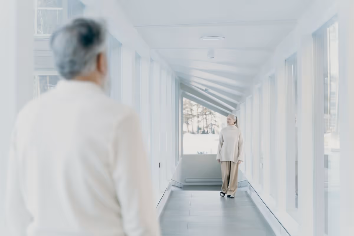 Two seniors stand in a bright, modern hallway with large windows at a senior living facility.