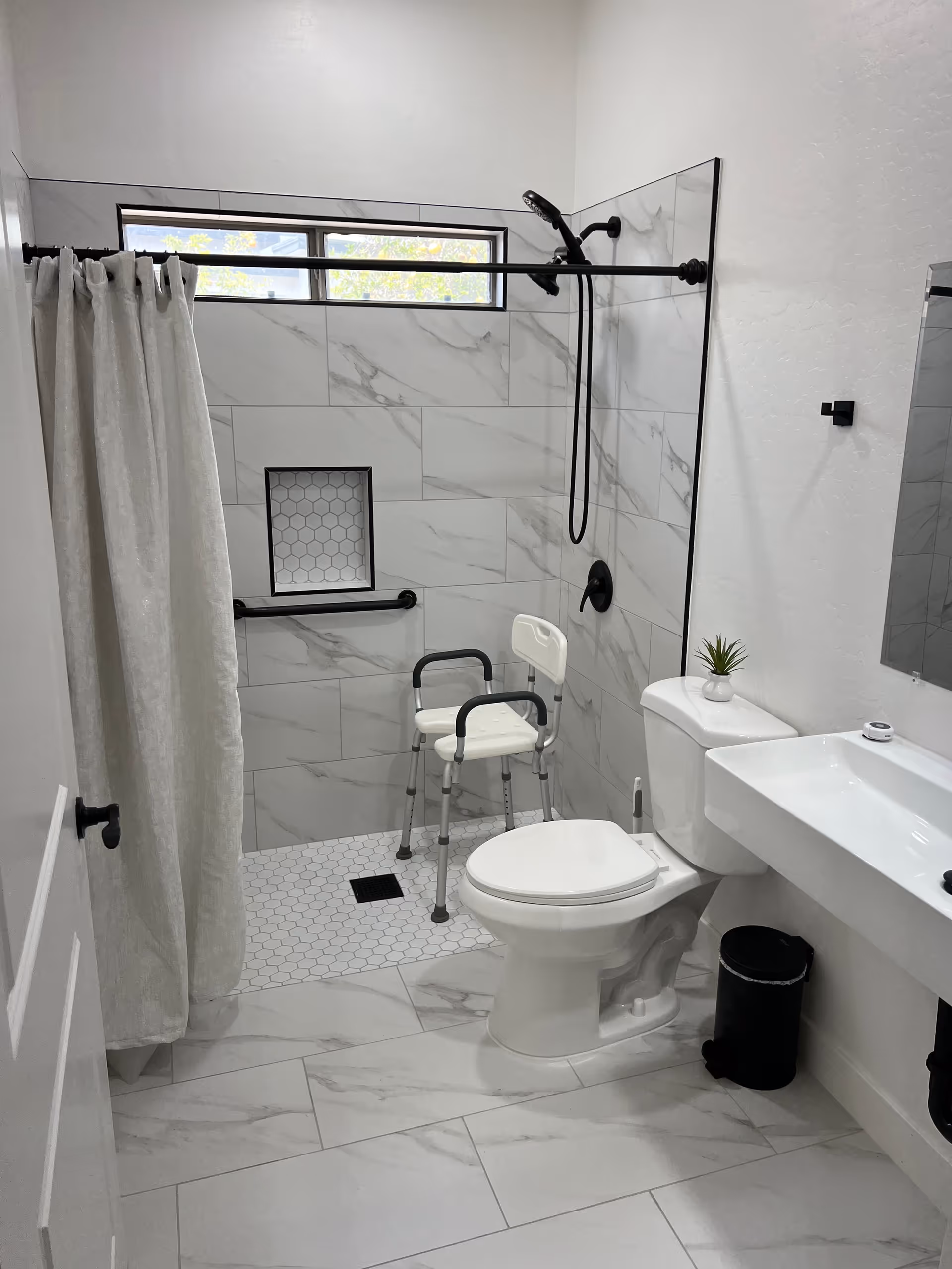 Accessible bathroom with shower chair, grab bars, curtain, toilet, sink, and plant.