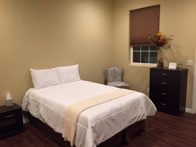 Neatly made bedroom with a bed, nightstands, dresser, a chair, and window blinds.