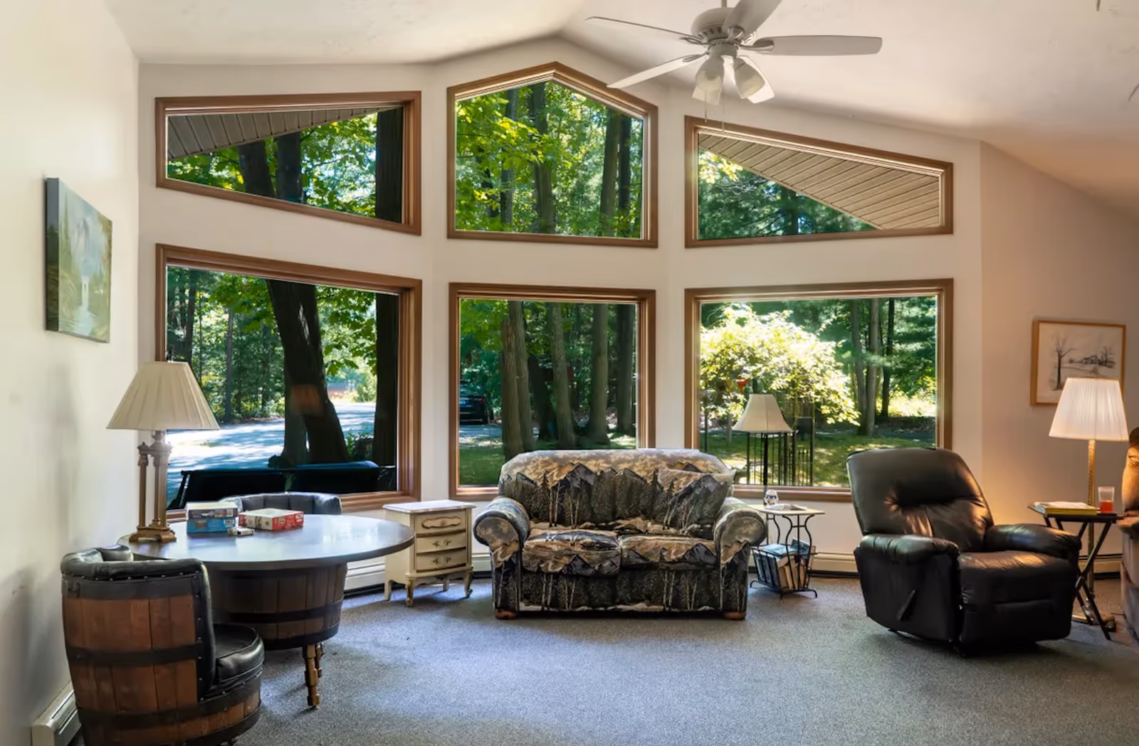 Bright living room with large window wall, sofa, recliner, and lamps overlooking trees.