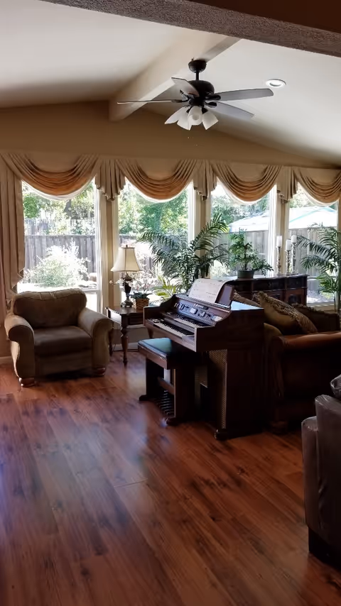 A cozy living room with wooden flooring, a ceiling fan, large windows with beige curtains, an armchair, a side table with a lamp, a piano with a bench, and several green plants. Outside the windows, a wooden fence and greenery are visible.