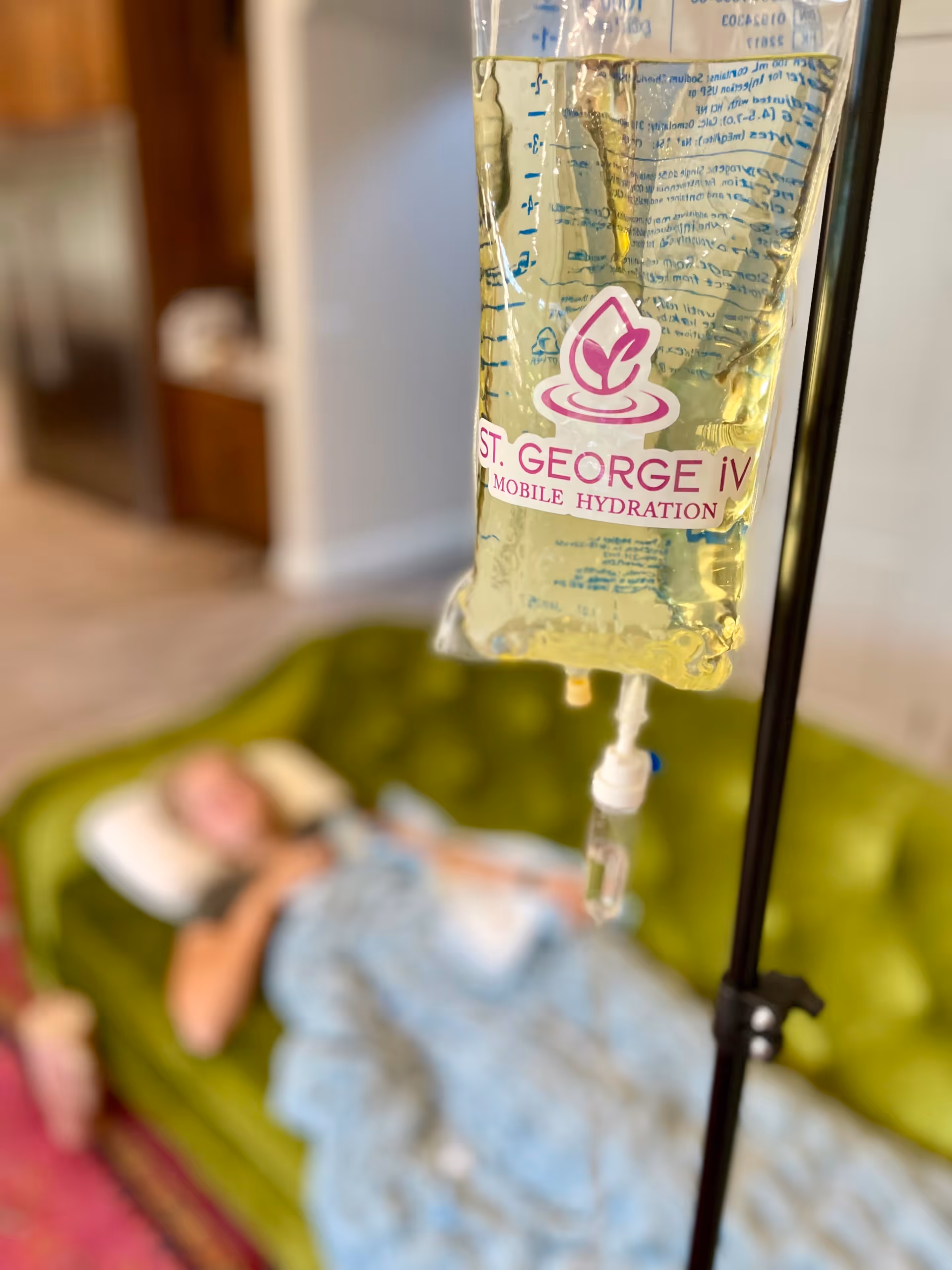 IV drip bag labeled St. George iV in foreground, patient resting on sofa in background.