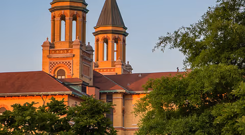 A red-brick building with two tall bell towers lit by warm light, partially framed by green trees in the foreground.