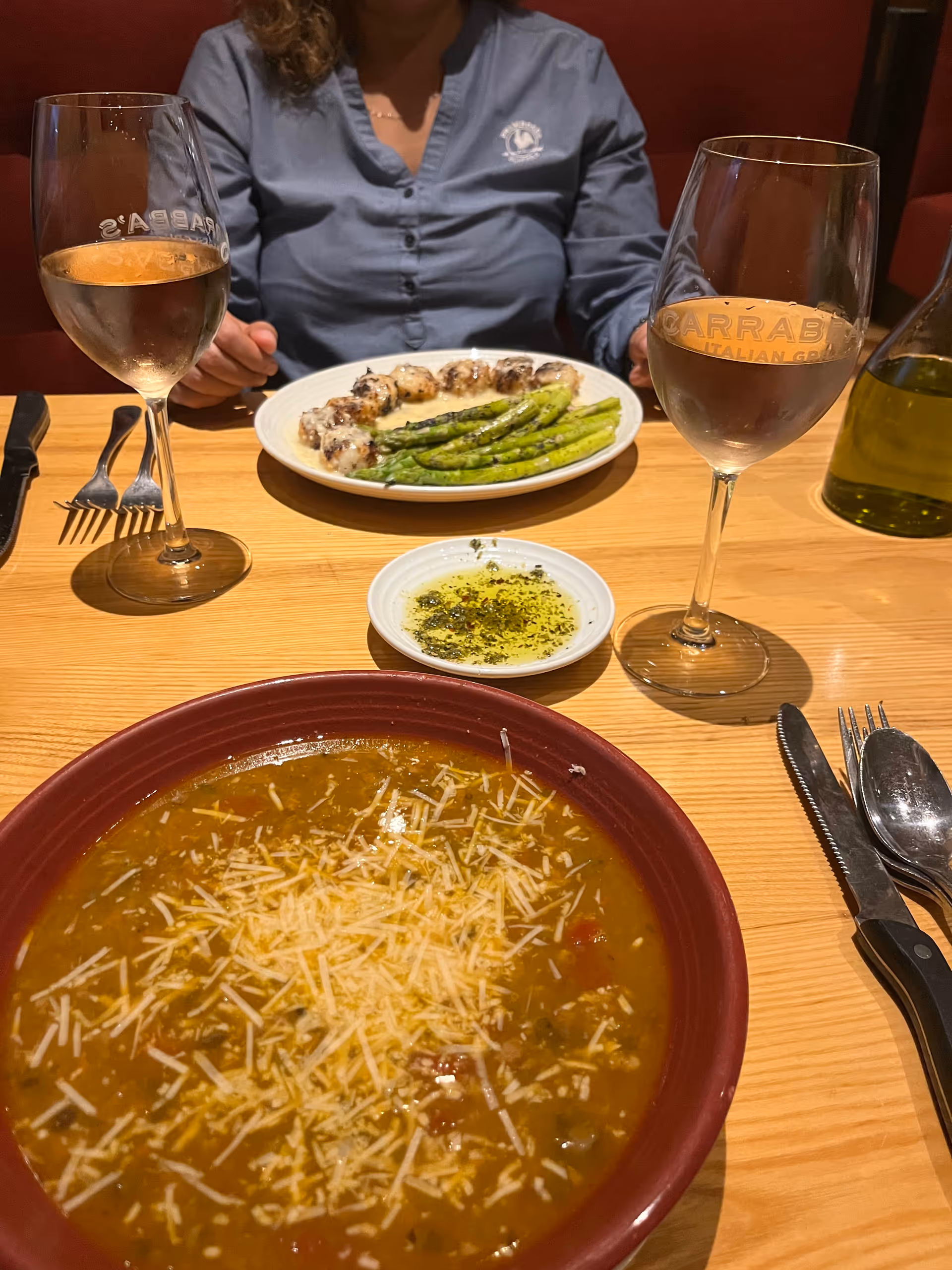 Person seated at a wooden dining table with soup, asparagus, and wine glasses.
