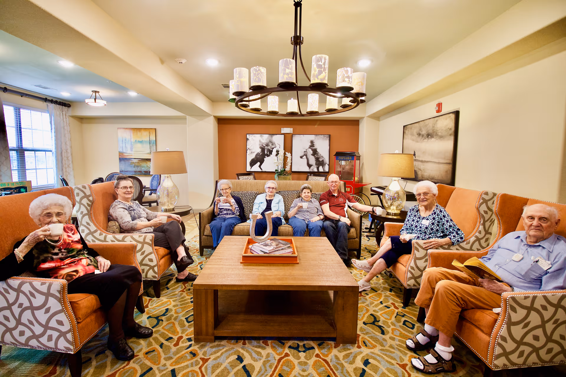 A group of elderly people sitting comfortably in a well-lit living room with orange and patterned armchairs and sofas around a wooden coffee table. The room has warm lighting from table lamps and a chandelier, with artwork on the walls and a popcorn machine in the background.