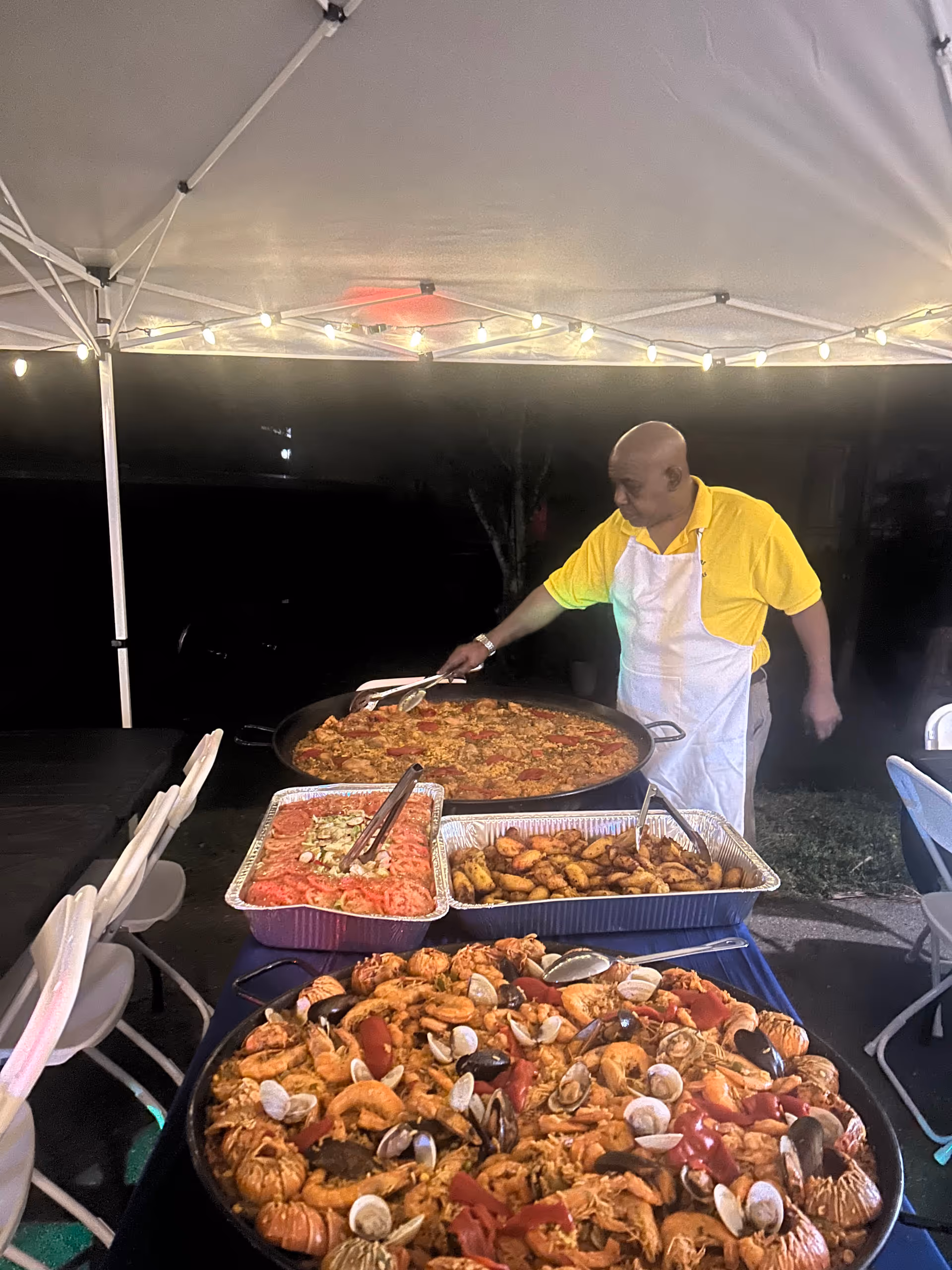 A staff member cooks paella beside seafood trays under string lights outdoors.