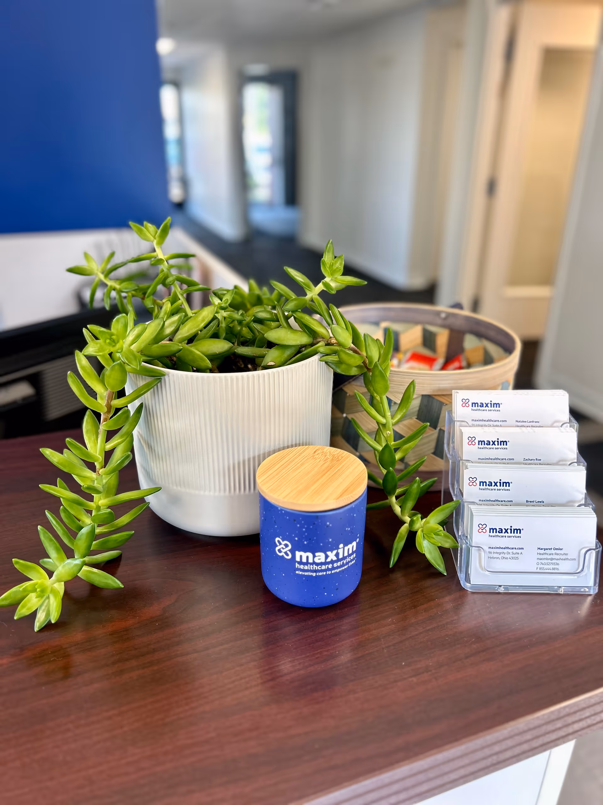 Reception desk with a potted plant and Maxim-branded items in a hallway.