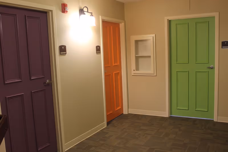 Hallway in a memory care facility showing three colorful doors (purple, orange, green), a wall sconce, and carpeted floor.