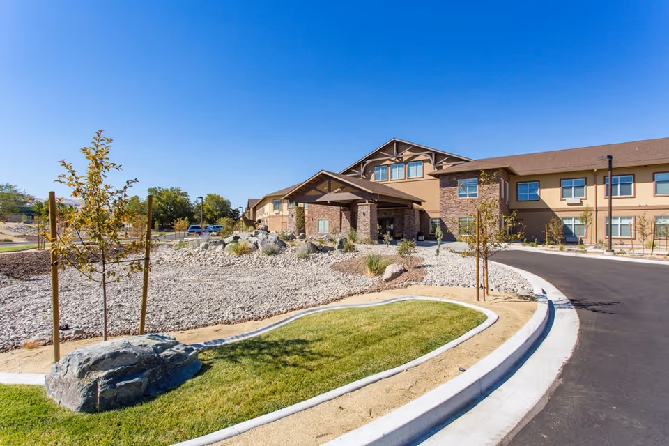 Front exterior of a two-story senior living building with a porte-cochère, landscaped rock garden and circular driveway under a clear blue sky.